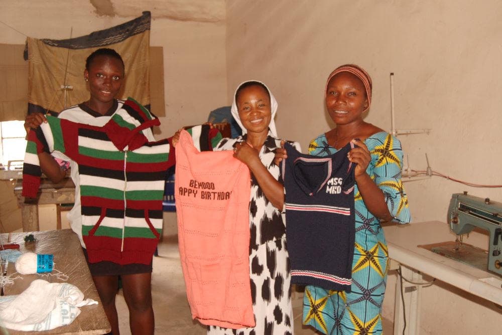 Three women smile as they hold up knitted sweaters they made, beside a sewing machine.