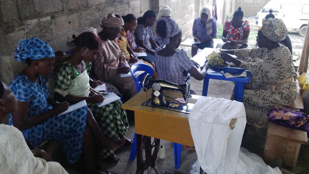 Women seated on benches take notes in exercise books beside a treadle sewing machine.